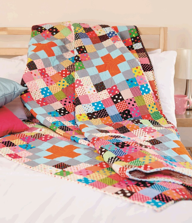 Multi-coloured patchwork quilt made of square blocks draped over a bed.