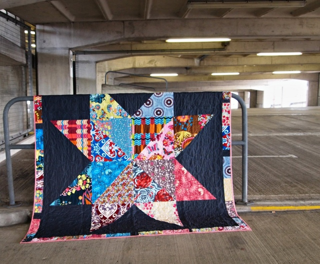 Large quilt with black background and large multi-coloured, patterned star shape in the centre with a multicoloured border, hanging over a railing in a carpark.