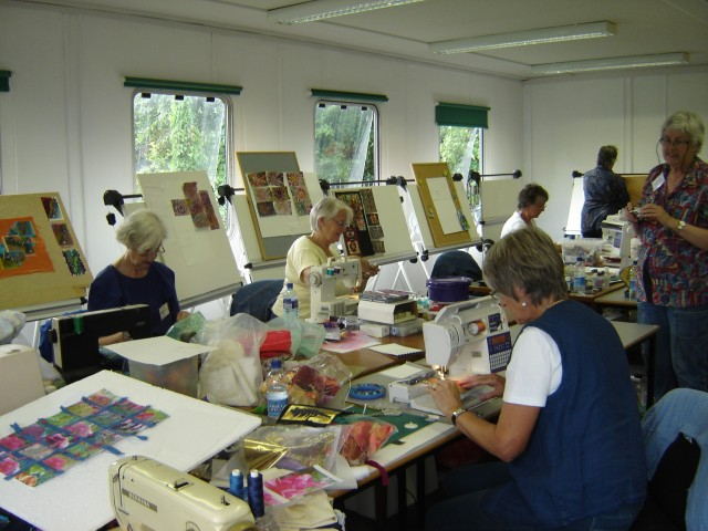 A group of ladies in a classroom, sat around a large table each working at a sewing a machine. Display boards are leaned up around the edge of the room.