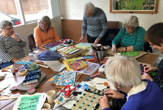 Group of women sat around a table on which there are piles of fabric and papers each working on a project.