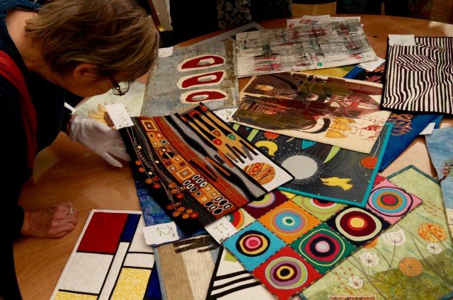 Woman, wearing a white glove, examining a selection of small quilts laid out on a table