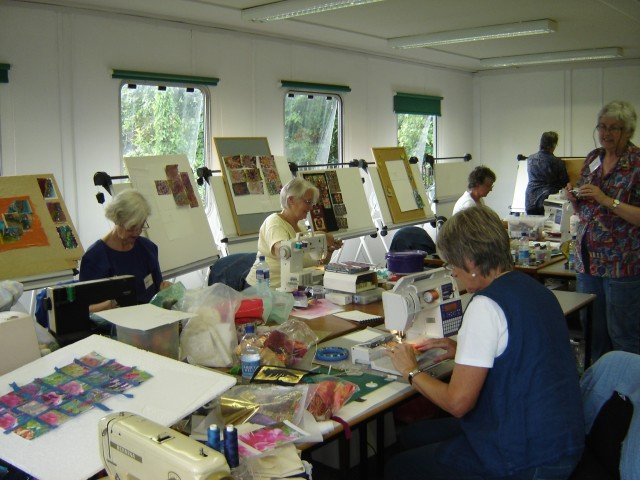 A group of ladies in a classroom, sat around a large table each working at a sewing a machine. Display boards are leaned up around the edge of the room.