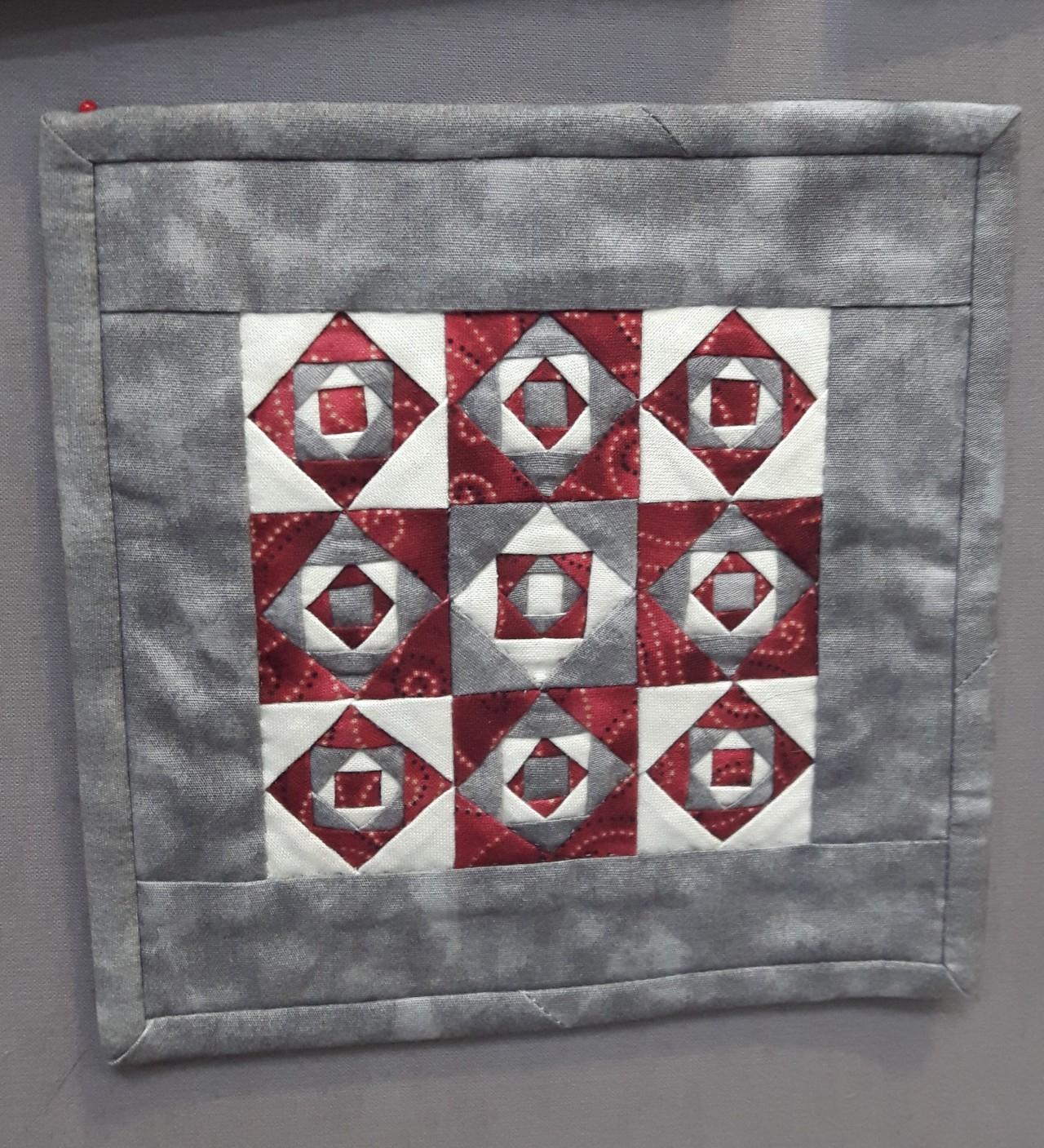 Square miniature quilt on display: grey background with a design of white and red squares in the centre