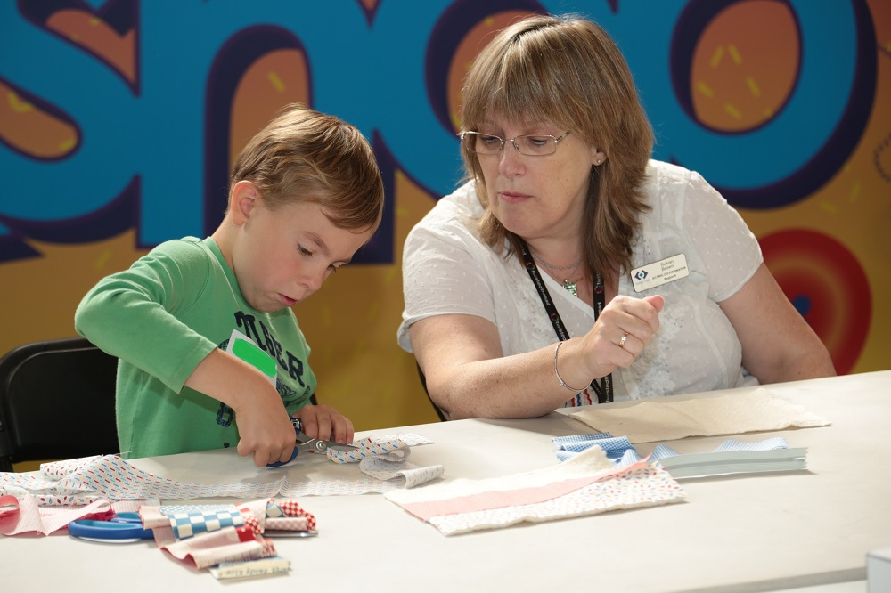 A woman and a boy sat at a craft table. The woman is supervising the boy cutting out fabric.