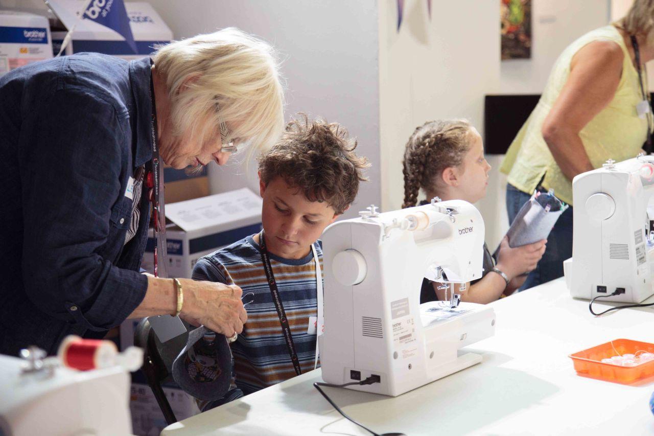 A women helping a boy, sat at a table with a sewing machine in front, with a sewing project