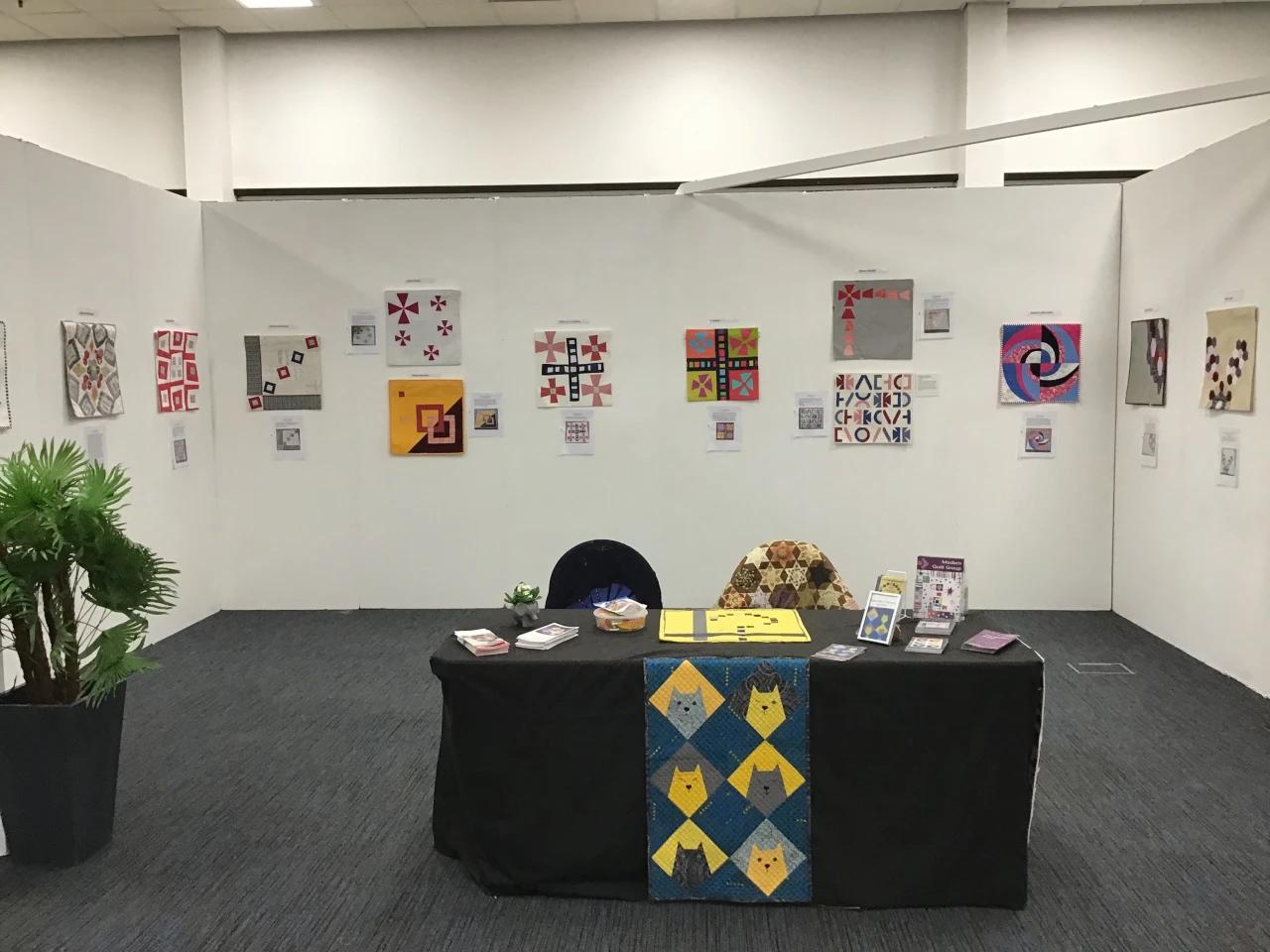 A table display of information leaflets in front of an exhibition wall displaying square shaped modern quilts