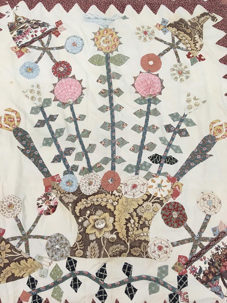 Appliqué top with a central motif of a basket with long stemmed flowers and four corner triangular baskets with star arrangement of flowers. This central panel is surrounded by frames of cable stems with flowers and leaves. The frame sections are partitioned by zigzag/triangular pieces.