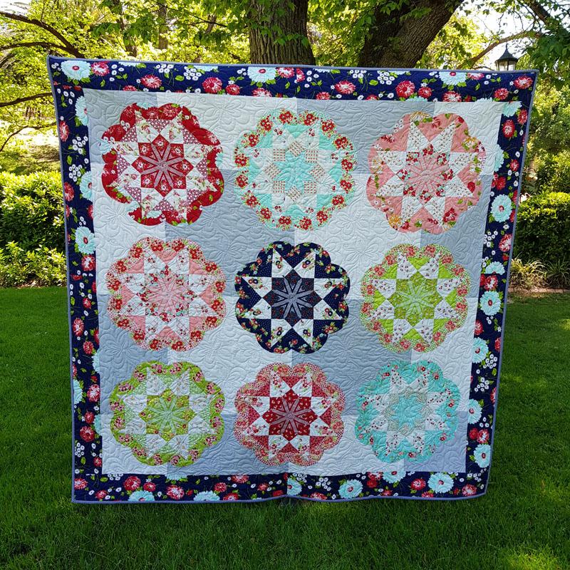 Large square quilt hung on line in a garden: Light blue and white checkerboard background with red, turquoise, green , pink and blue repeated flower pattern and a navy blue border with light blue and red flowers