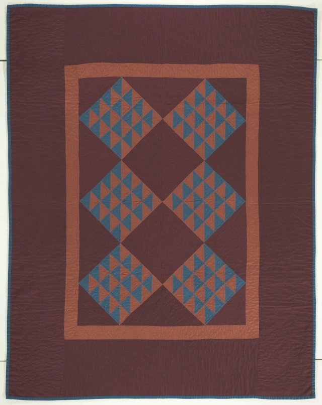 Large rectangular burgundy quilt with a muted orange rectangle in the middle. Within which, is 6 diamond shapes made up small blue and muted orange triangles