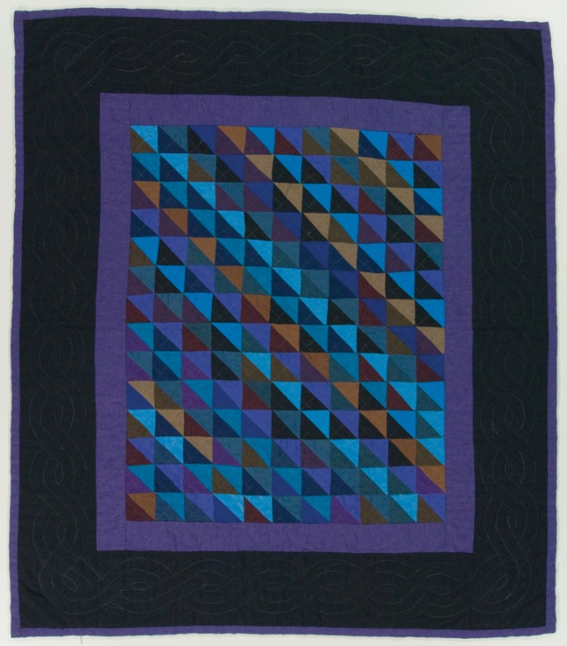 Rectangular quilt with wide black border with a purple edge. The centre design is made of a purple border with a repeated pattern of purple, beige, blue, burgundy and orange triangles in the middle.