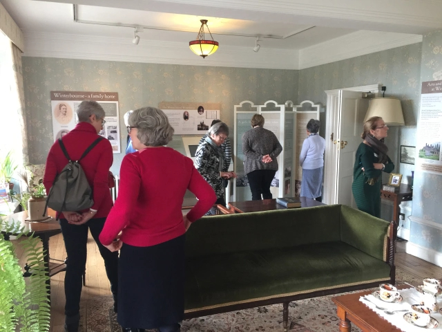 Guild members explore a room in the house with a dark green sofa, a coffee table set for tea and its interpretation displays.