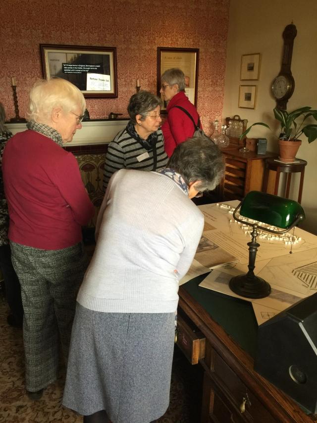 Guild members peruse John Nettlefold?s study: A large desk with a desk lamp and papers on top. A plant on a plant stand in the corner