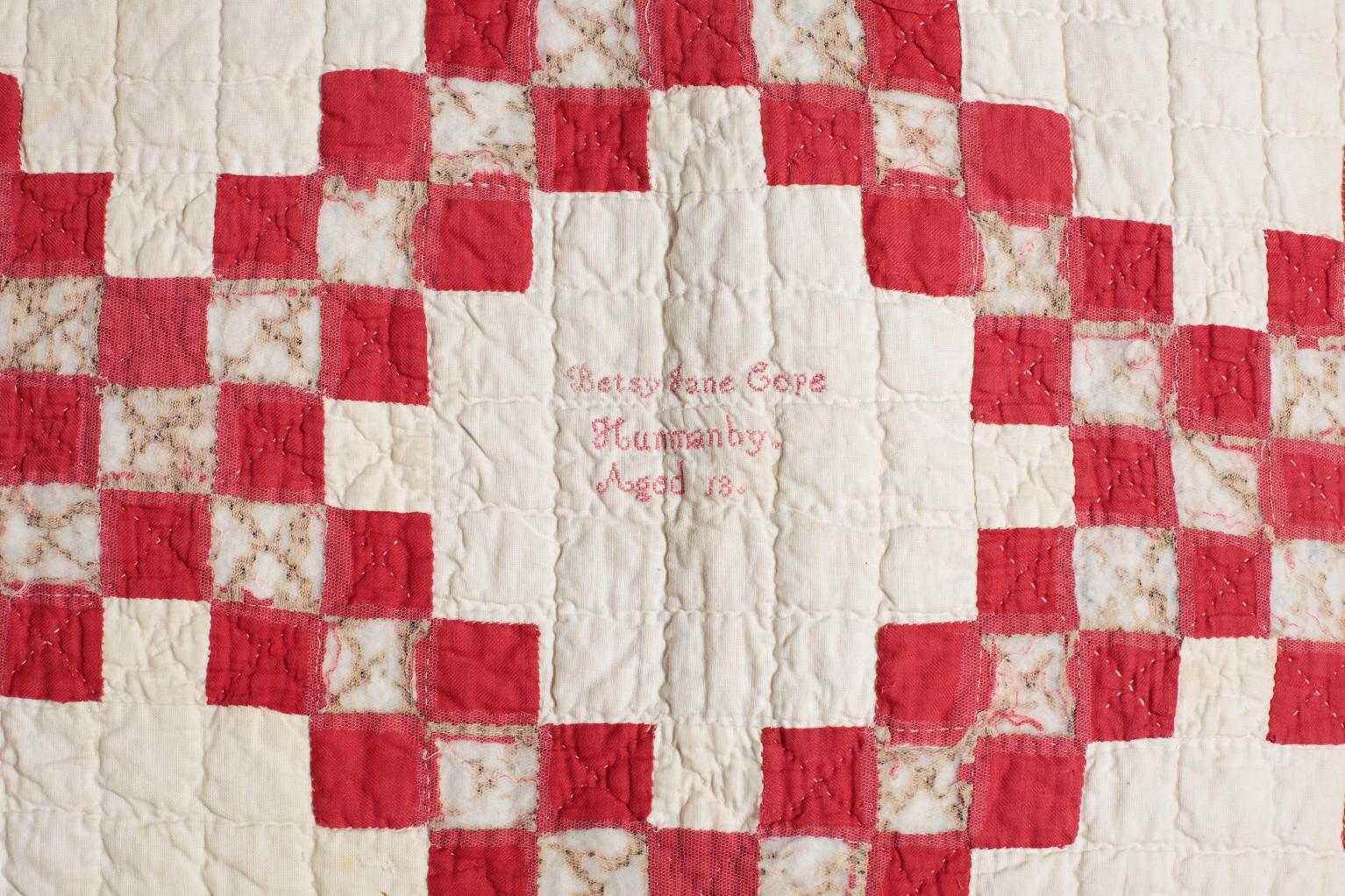Close up of the Betsy Cope quilt: Red squares on a white background with a embroidered text saying 'Betsy Jane Cope, Hunmanby, Aged 13'
