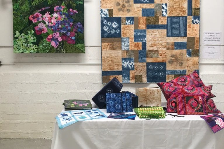 A table displaying quilt examples at an exhibition in front of two quilts hung on display on a wall