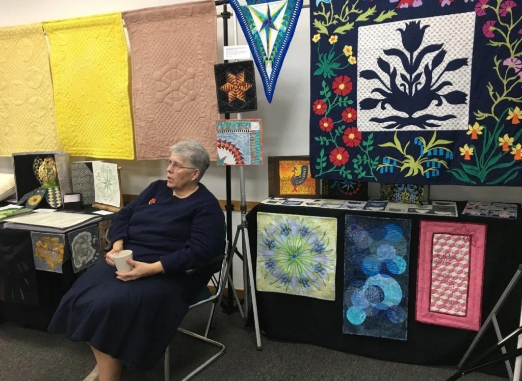 Jackie Benstead sat on a chair in front of her quilts on display at the Festival of Quilts