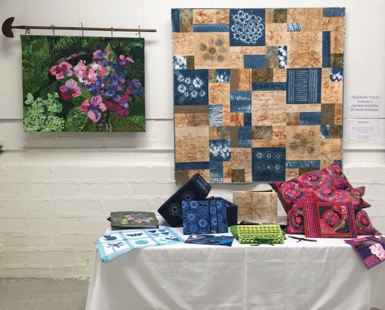 A table displaying quilt examples at an exhibition in front of two quilts hung on display on a wall