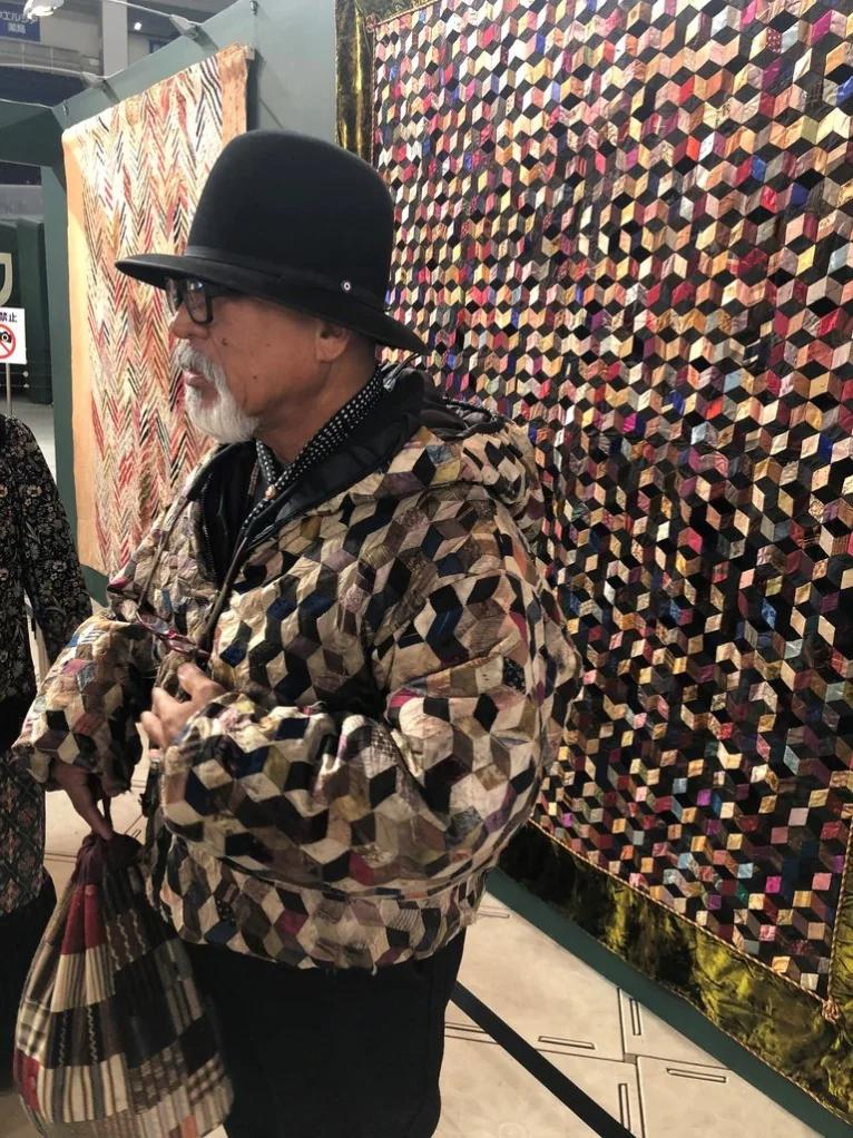 Man stood in front of a multicoloured quilt on display wearing a quilted jacket of a similar design