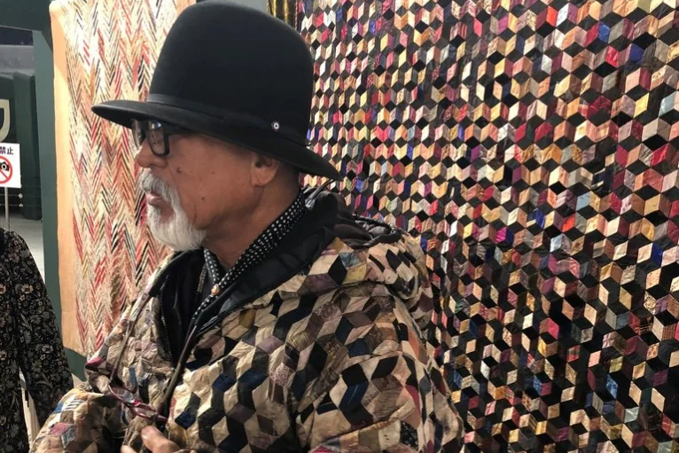 Man stood in front of a multicoloured quilt on display wearing a quilted jacket of a similar design