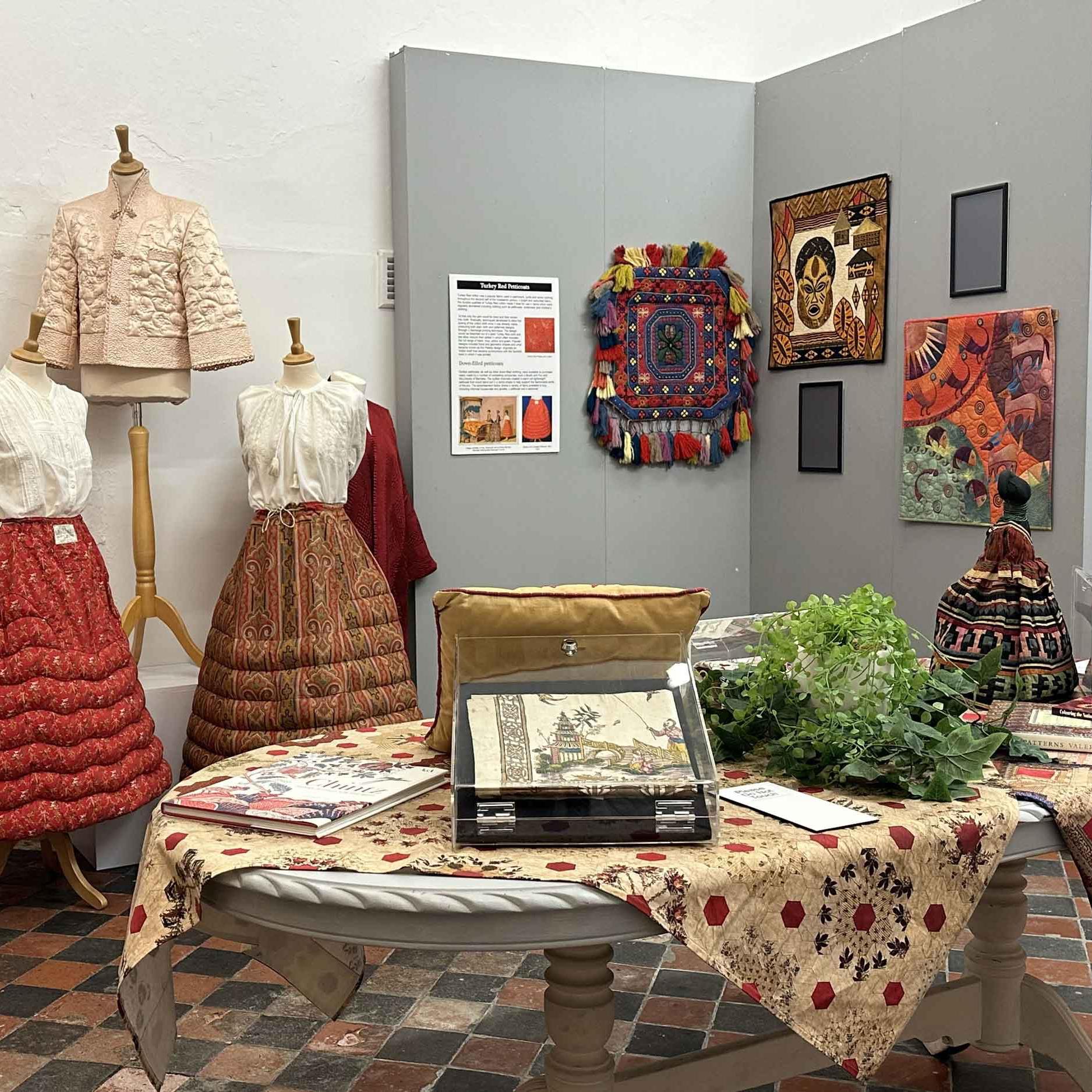 An image of a gallery room with a range of internationally inspired quilted items including quilted petticoats, a quilted smoking jacket and a hand-stitched doll.