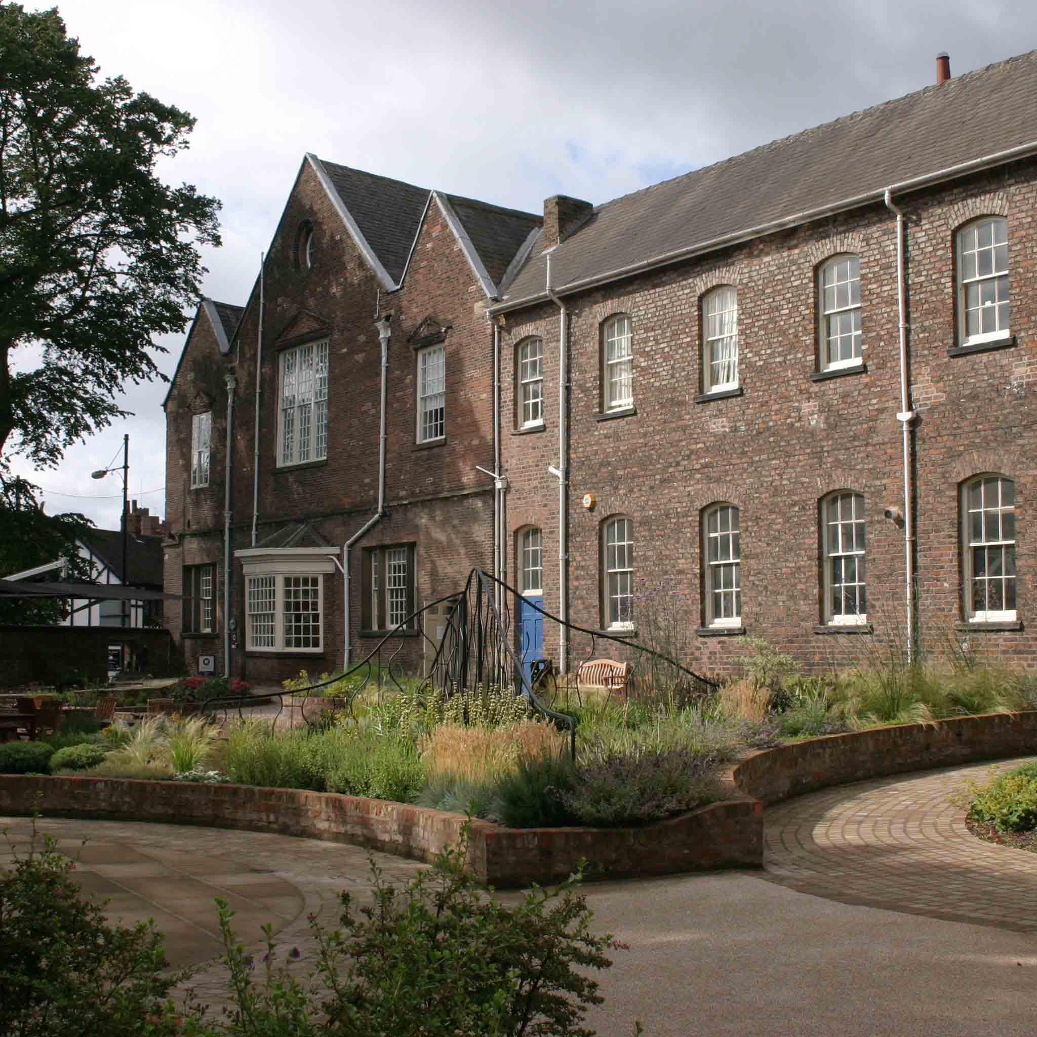 An image of a grand brick building with a short walled sensory garden with plants and decorative structures