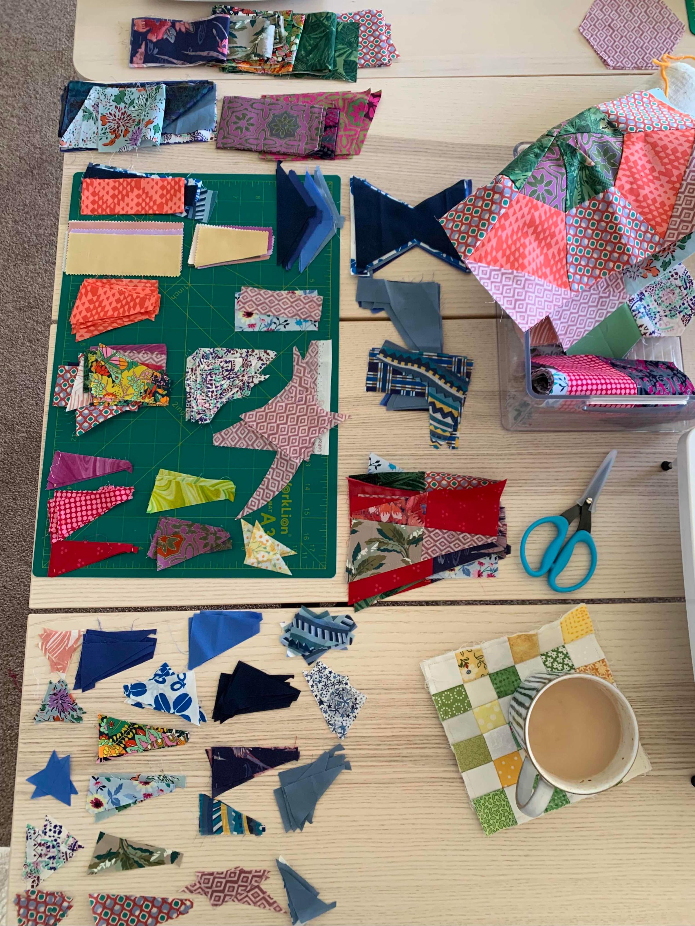 A table covered in bright fabric scraps, scissors and a cup of tea