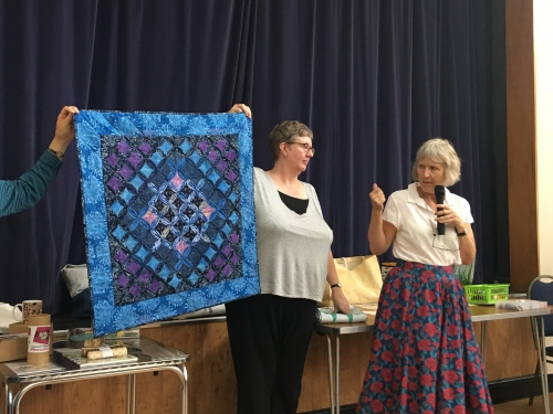Woman with microphone speaking, whilst stood next to woman holding up blue and purple patterned quilt