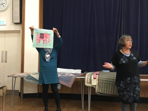 2 women standing in front of crowd. 1 holding up a quilt with a pink elephant on a turquoise background
