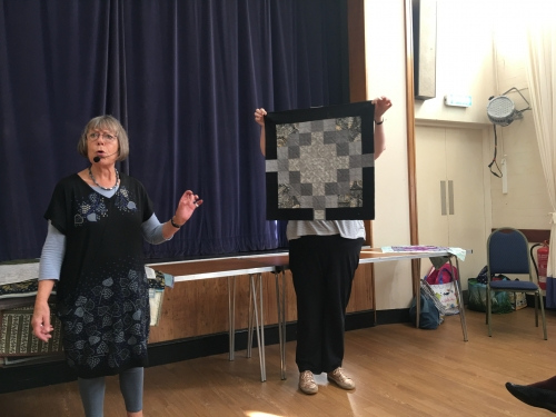 2 women standing in front of crowd. 1 holding up a quilt with square patterns.