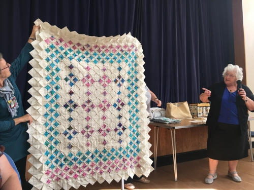 Woman with microphone speaking, whilst stood next to people holding up large quilt with triangle repeated triangle shaped edge and a beige, blue and pink pattern