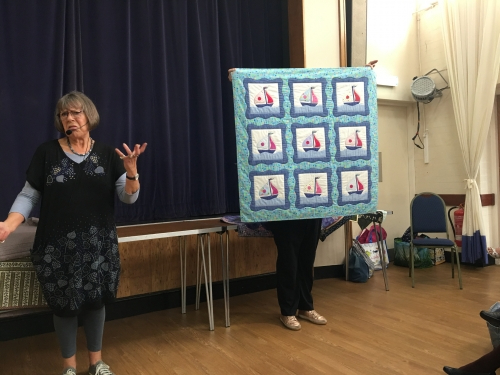 2 women standing in front of crowd. 1 holding up a quilt made of 9 blocks showing a sailing boat with a blue border.
