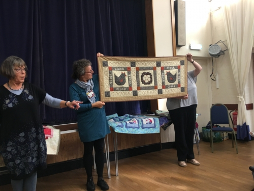 2 women standing in front of crowd. 1 holding up a long quilt with chicken and egg patterns