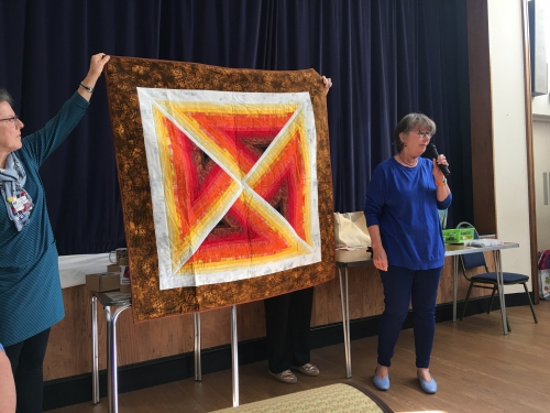 Woman with microphone speaking, whilst stood next to people holding up quilt with brown border and yellow and red triangle centre