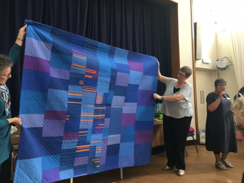 Woman with microphone speaking, whilst stood next to woman holding up blue and purple block patterned quilt