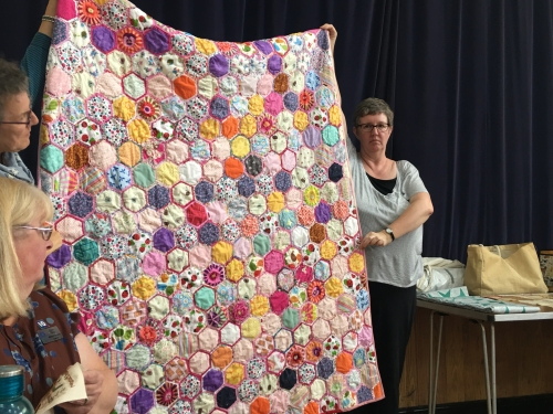 Women holding up large quilt made of multi-coloured hexagonal pieces