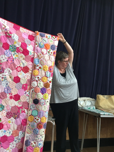 Women holding up large quilt made of multi-coloured hexagonal pieces showing pink back
