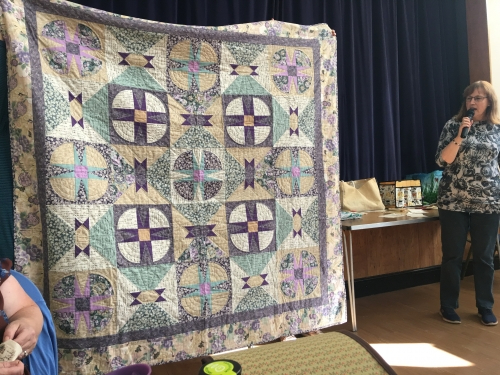 Woman with microphone speaking, whilst stood next to people holding up patchwork quilt with circles and diamonds in greens, yellows and purples