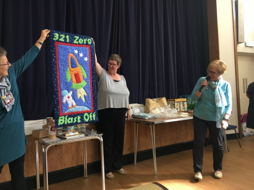Woman with microphone speaking, whilst stood next to people holding up quilt with rocket and astronaut in centre