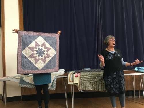 2 women standing in front of crowd. 1 holding up a quilt with floral design.