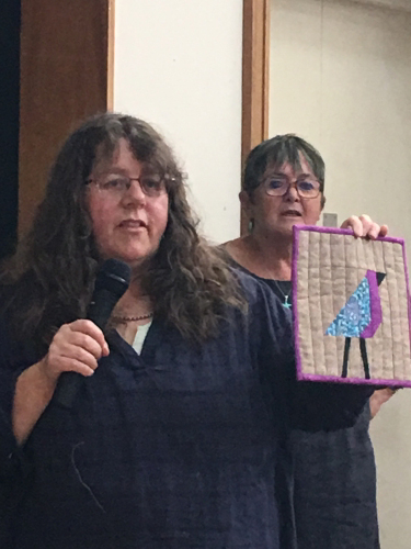 Two women. One holding a microphone in one hand and a miniature quilt with pink border and bird design