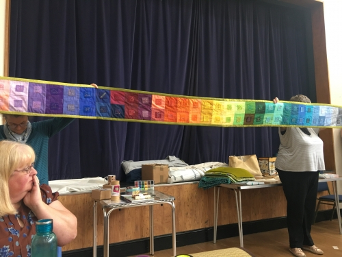 People holding middle of very long and narrow quilt with multicoloured square designs