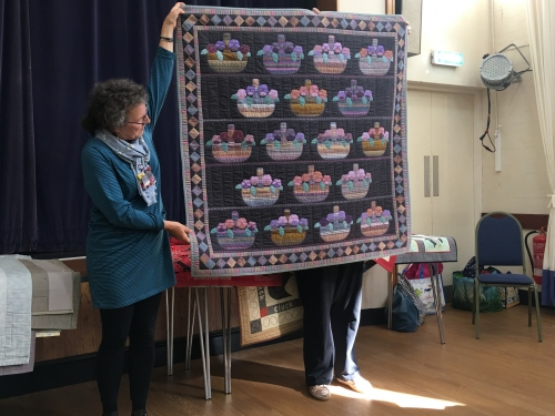 Woman holding up quilt with black background, purple border and multiple baskets of pink and purple flowers