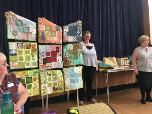 Woman talking into microphone, stood in front of people holding large multicoloured quilt made of 9 sections joined by cords