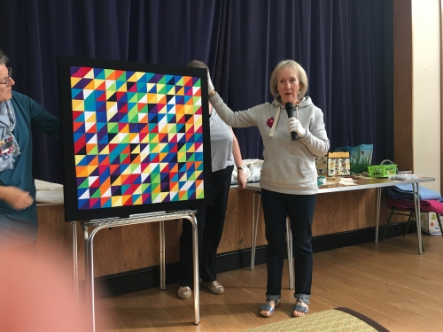 Woman with microphone speaking, whilst stood next to people holding up multi-coloured triangle patterned quilt