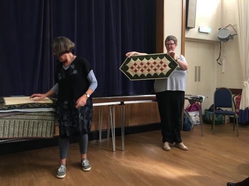 2 women standing in front of crowd. 1 holding up a quilt with diamond patterns.