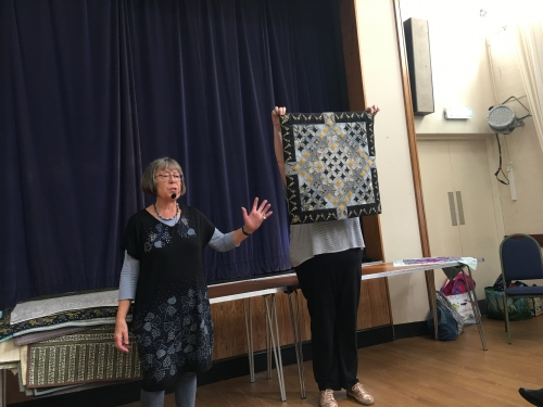 2 women standing in front of crowd. 1 holding up a quilt with diamond patterns.