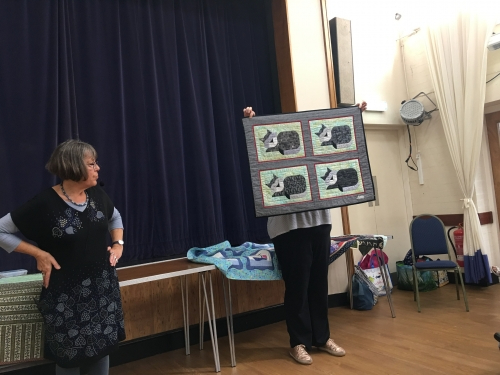 2 women standing in front of crowd. 1 holding up a quilt with cat patterns.
