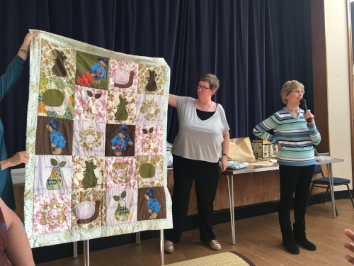 Woman with microphone speaking, whilst stood next to woman holding up quilt made of square blocks with flowers, pears and Paddington Bear