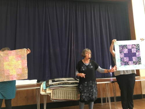 Woman speaking to crowd with microphone, with two people holding up quilts