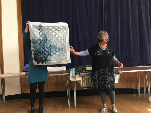 2 women standing in front of crowd. 1 holding up a quilt with geometric patterns.