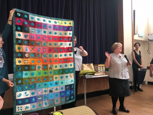 Woman talking into microphone, stood in front of people holding large quilt with multi-coloured squares and circles design